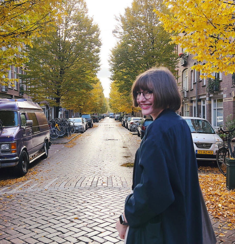A woman in a navy blue coat and glasses looks over her shoulder and smiles at the camera. She is standing on a brick laid street in Amsterdam.